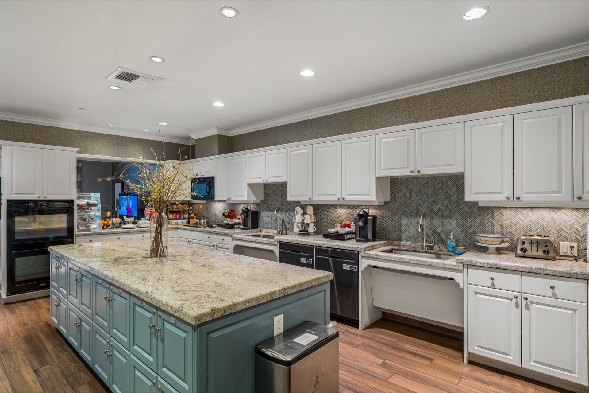 Large community kitchen with a granite island, white cabinetry, double ovens, and multiple sinks and cooking stations.