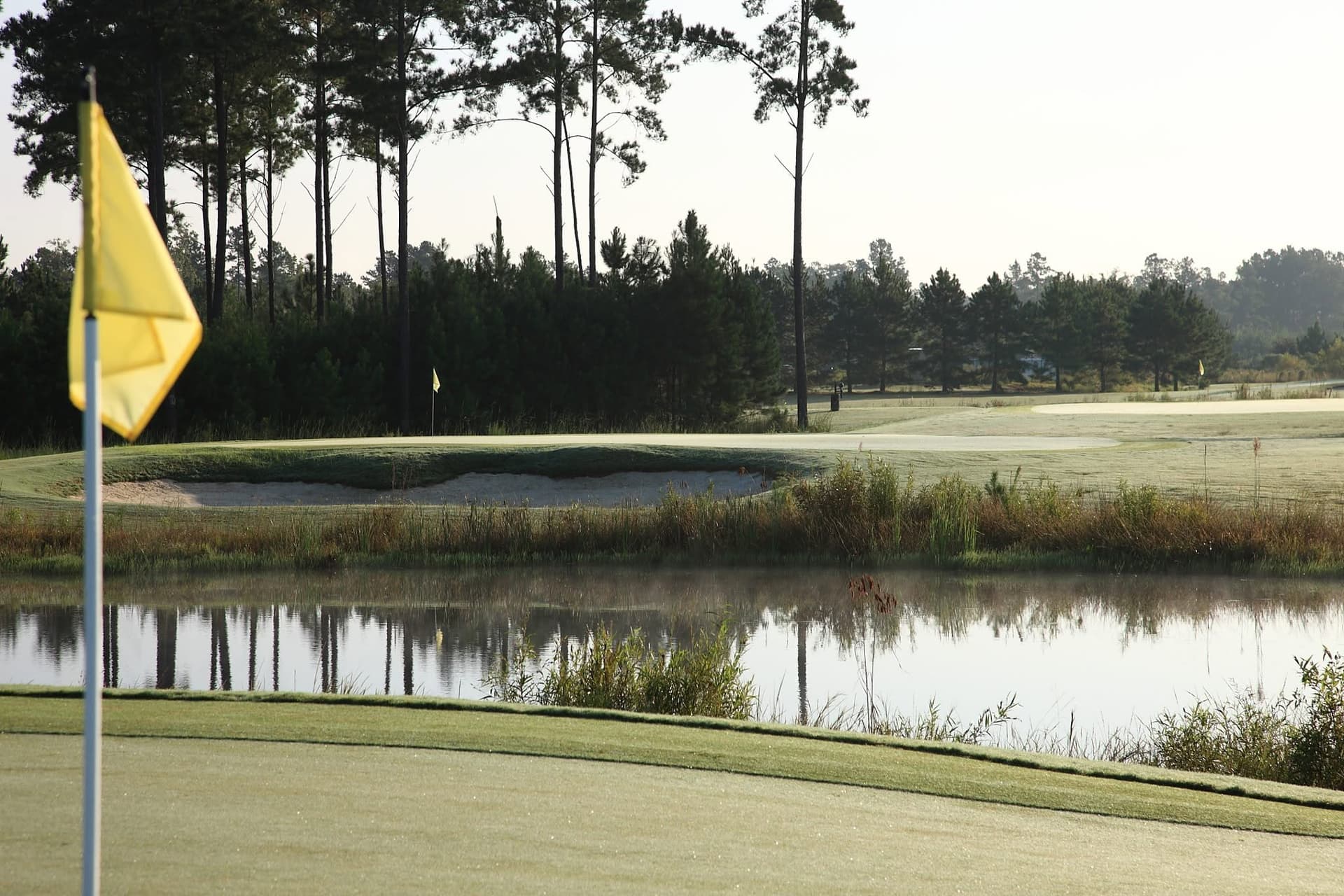 Morning light over a yellow flag, water hazard, and green at The Golf Club at Hilton Head Lakes, with tall pines in the background