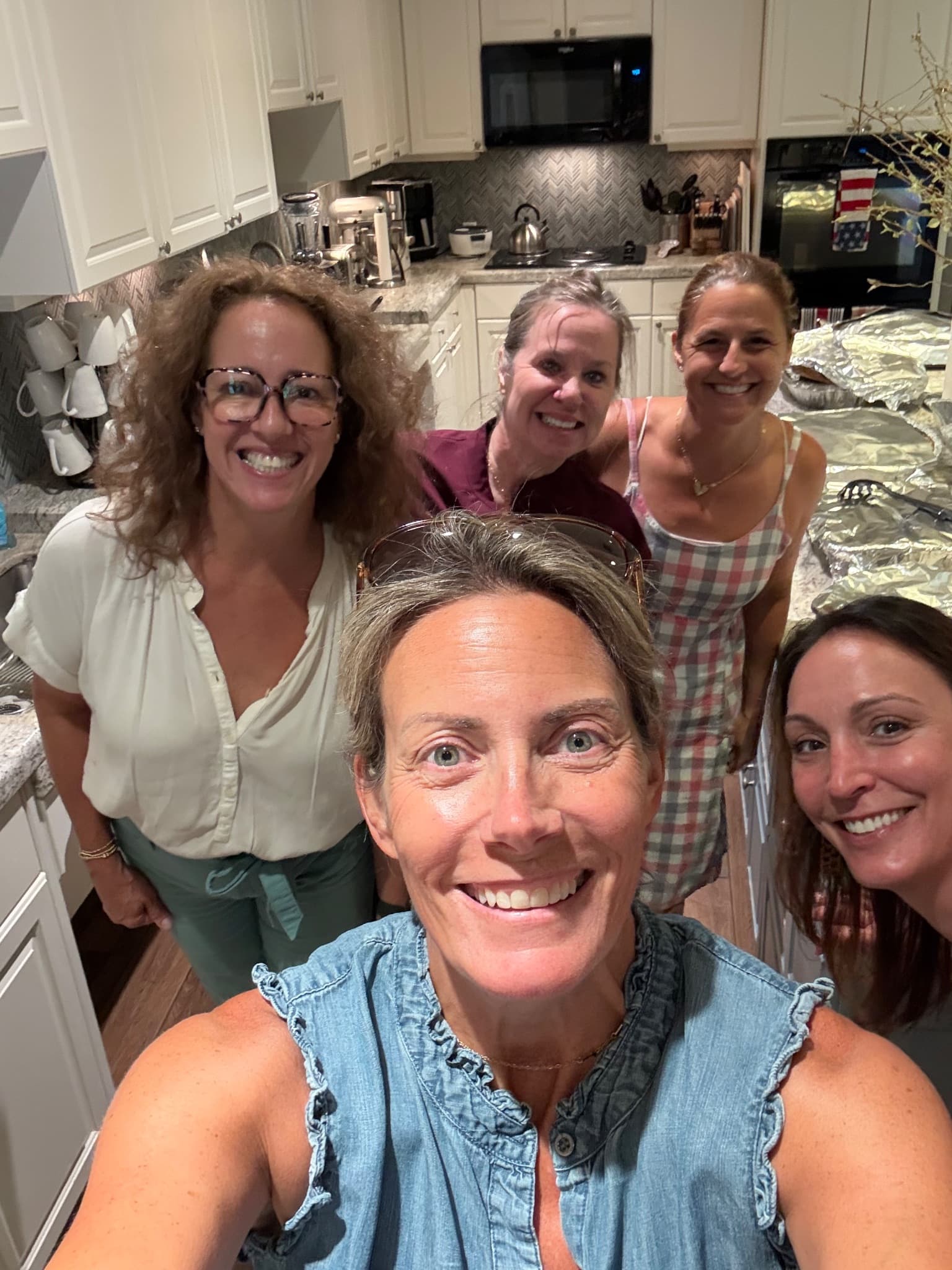 Five Anderson Bookclub volunteers taking a selfie in the Fisher House kitchen, with foil trays of pasta dinner lined up on the counter behind them