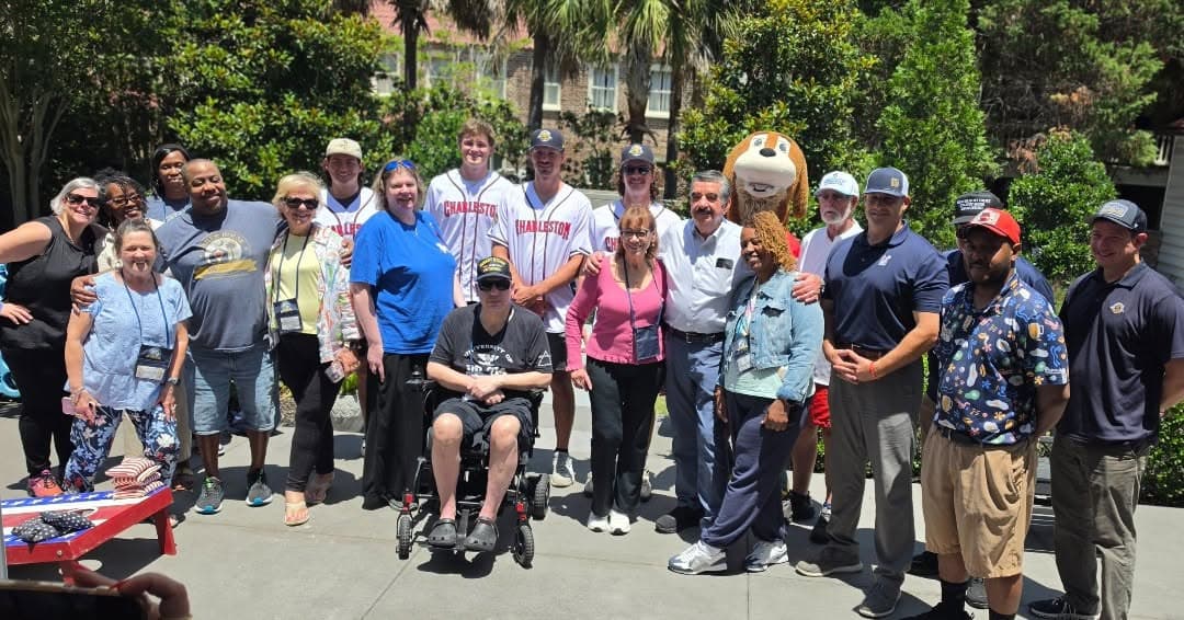 Charleston RiverDogs players in white jerseys, the Charlie T. RiverDog mascot, veteran families, and volunteers gathered together outdoors at the Charleston Fisher House