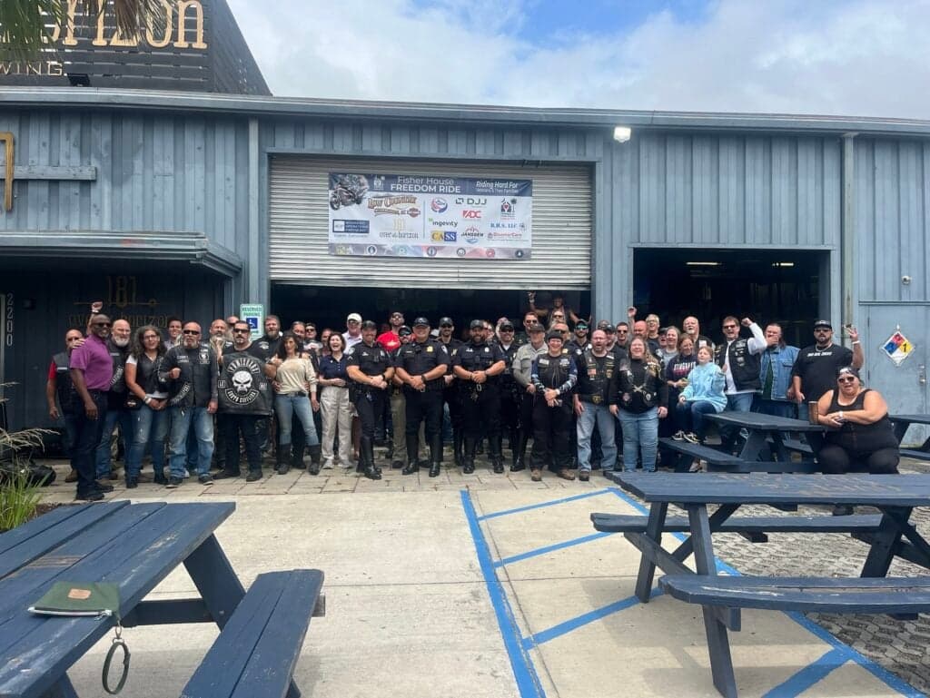 Riders, law enforcement officers, and volunteers gathered in front of the Fisher House Freedom Ride sponsor banner outside Over the Horizon Brewing
