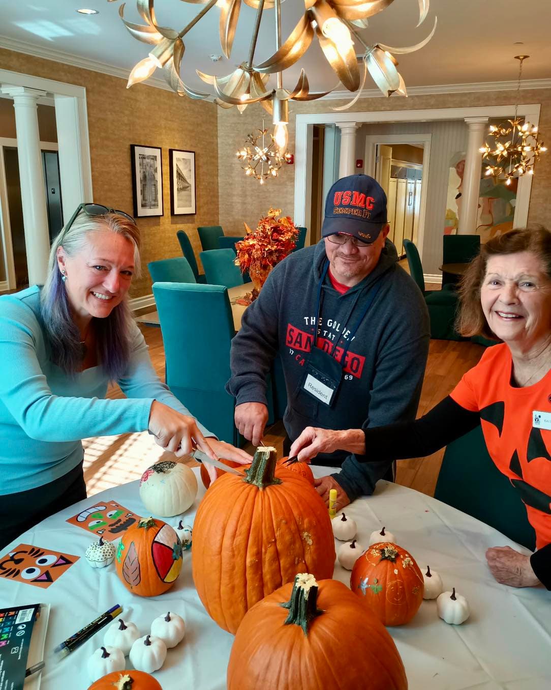 Two volunteers and a Fisher House guest in a USMC cap carving and decorating pumpkins together at a table in the Fisher House common room