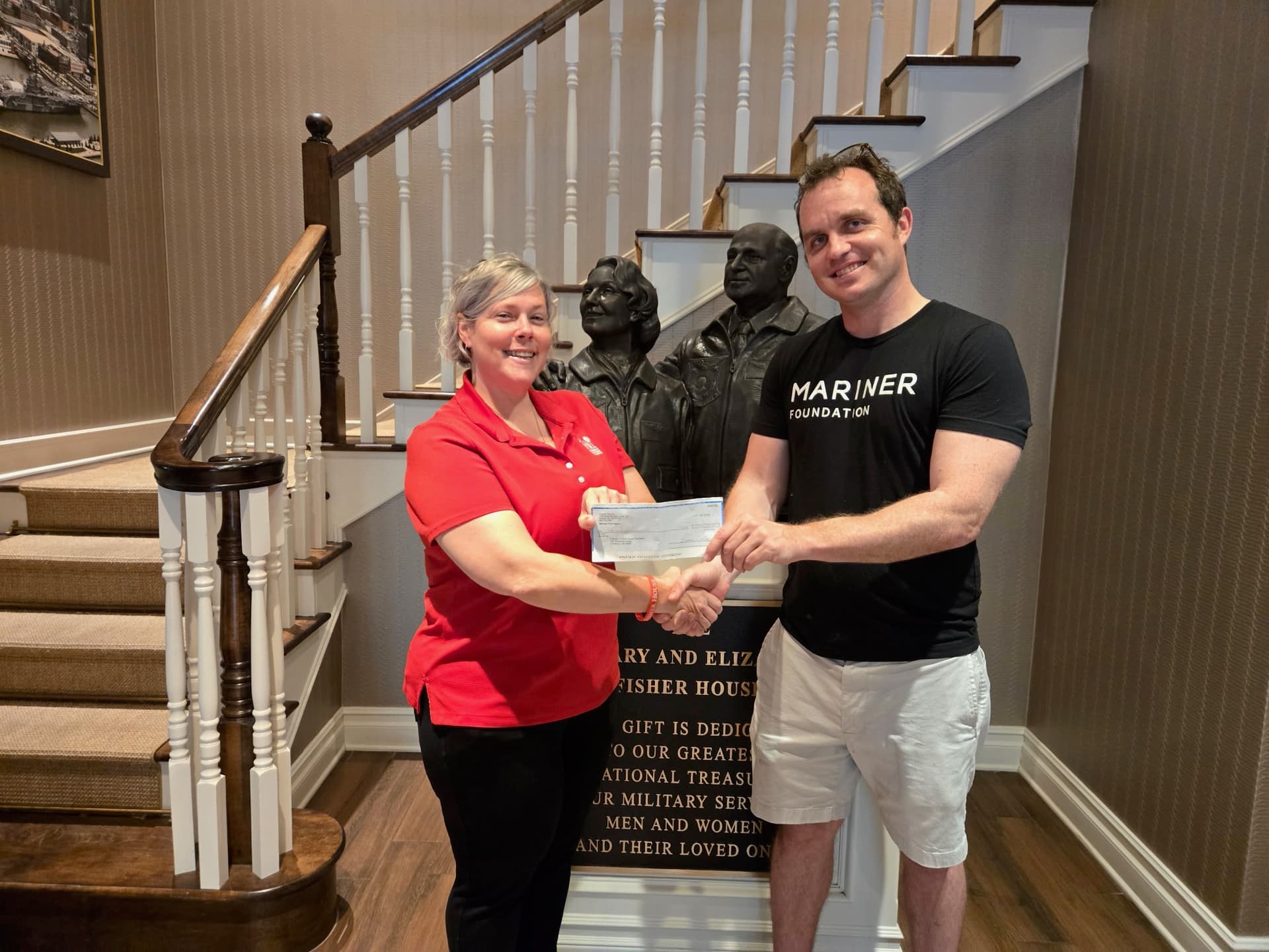 A Friends of Fisher House Charleston staff member in a red polo and a Mariner Foundation team member shaking hands while holding a $2,500 check in front of the Fisher House bronze sculpture and dedication plaque