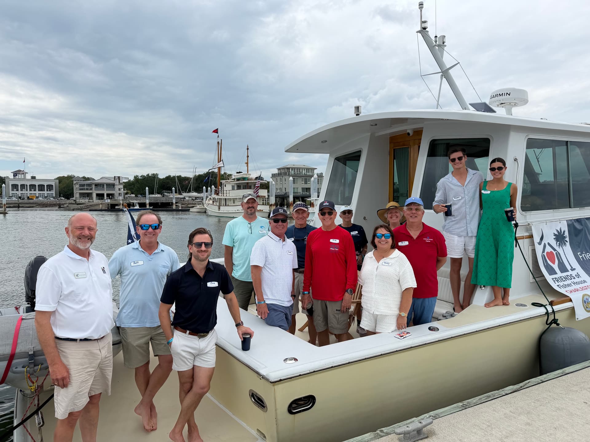 Friends of Fisher House Charleston supporters and Military Salute Regatta participants posing together on the stern of a yacht at the marina, with sailboats and Charleston Harbor in the background