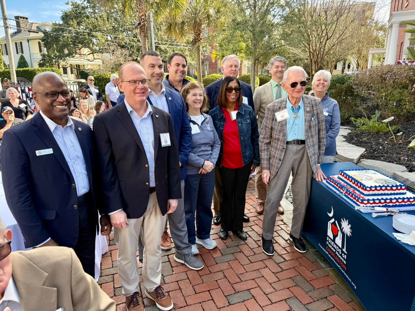 Supporters and staff gathered outside at the Friends of Fisher House Charleston open house, with a decorated cake on a branded table