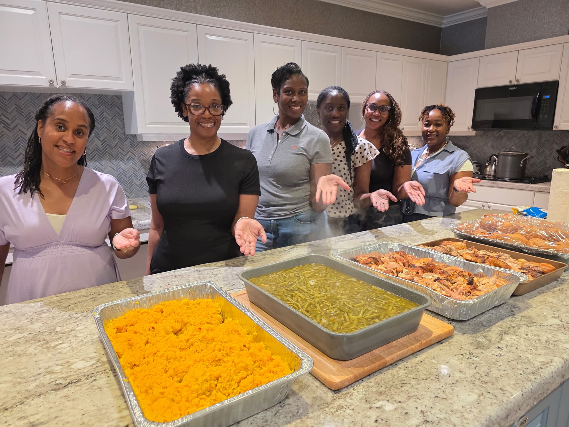 Six Royal Missionary Baptist Church YWA volunteers smiling behind the Fisher House kitchen island, with trays of yellow rice, string beans, and chicken in the foreground