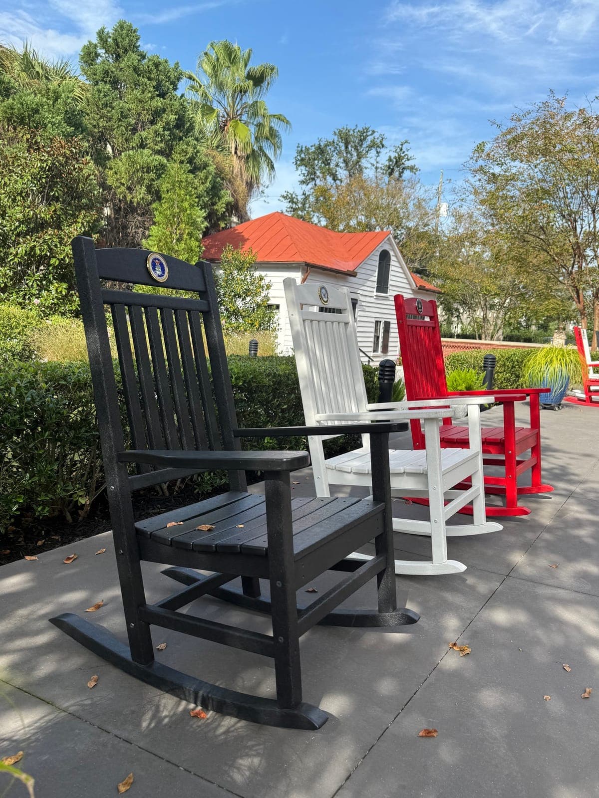 Black, white, and red rocking chairs on the Fisher House patio, each displaying a military service branch emblem, with the carriage house in the background