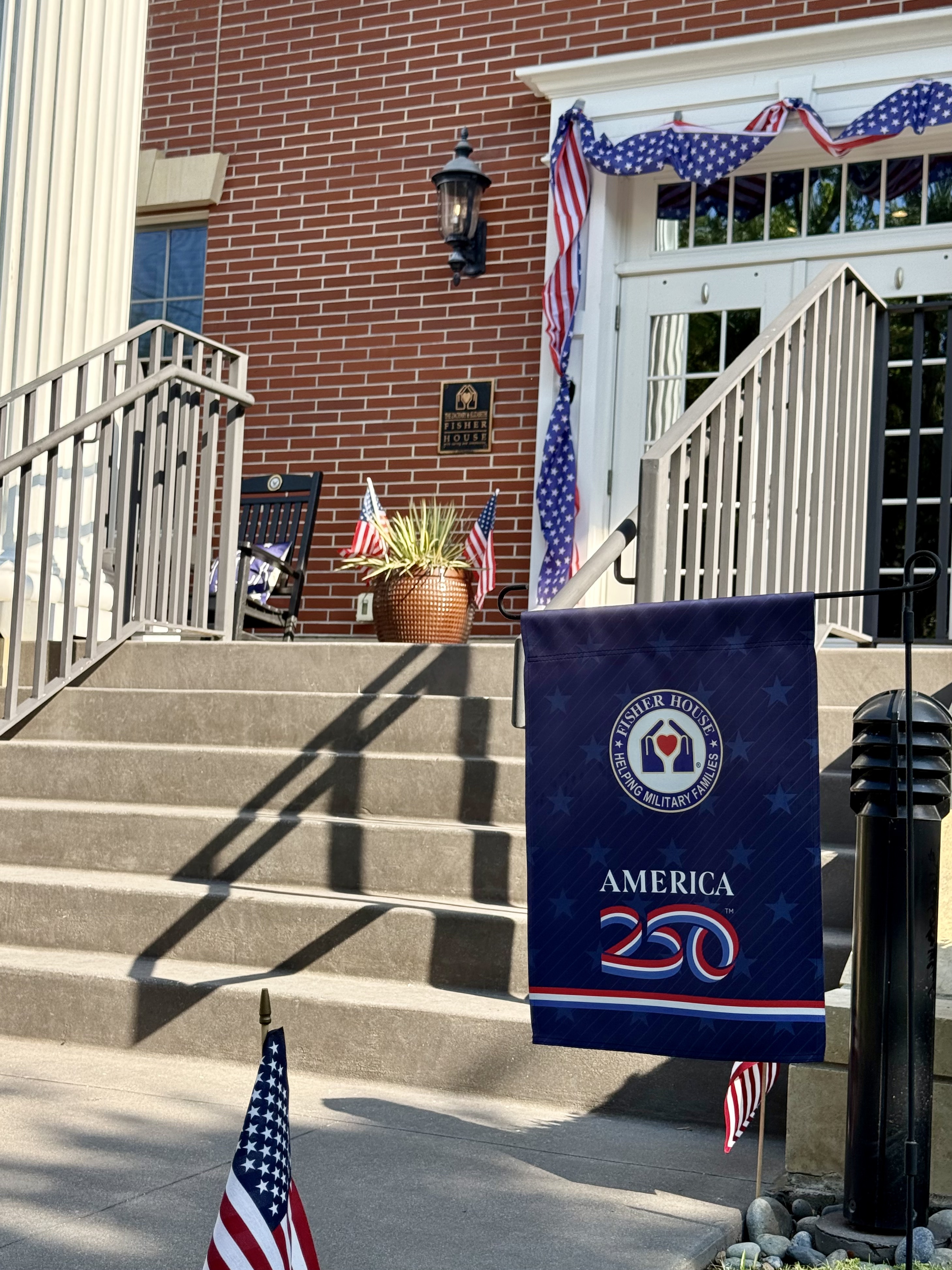 The front steps of the Charleston Fisher House decorated with patriotic bunting and American flags, with a Fisher House America 250 garden flag in the foreground
